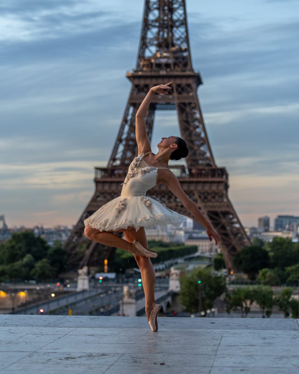 A ballerina in a white tutu on pointe, one arm raised, on the empty Trocadéro terrace with the Eiffel Tower directly behind her at dawn.
