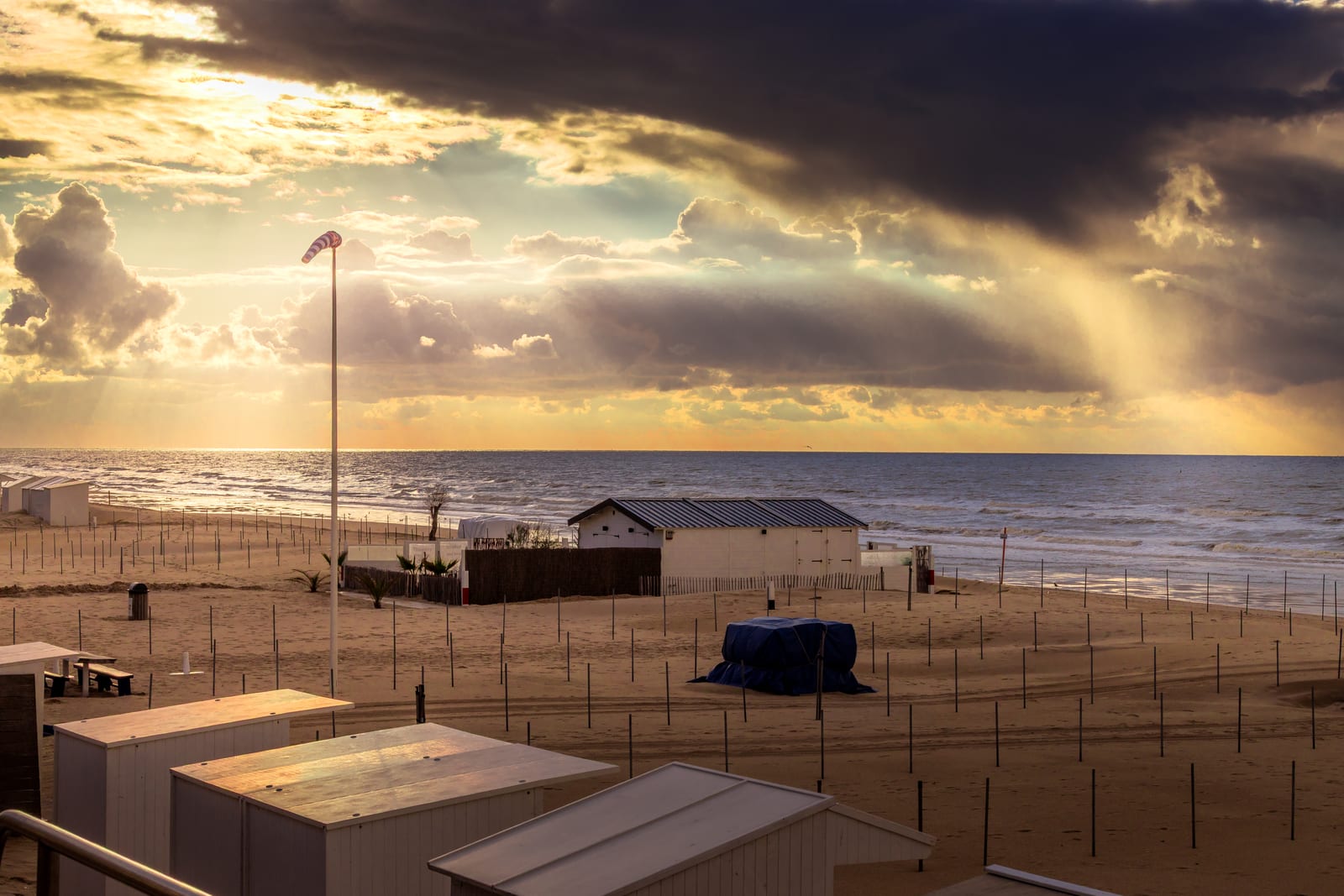 Beach huts and windbreaks on the sand at De Haan, with dark storm clouds over the North Sea and sunrays falling on the water.
