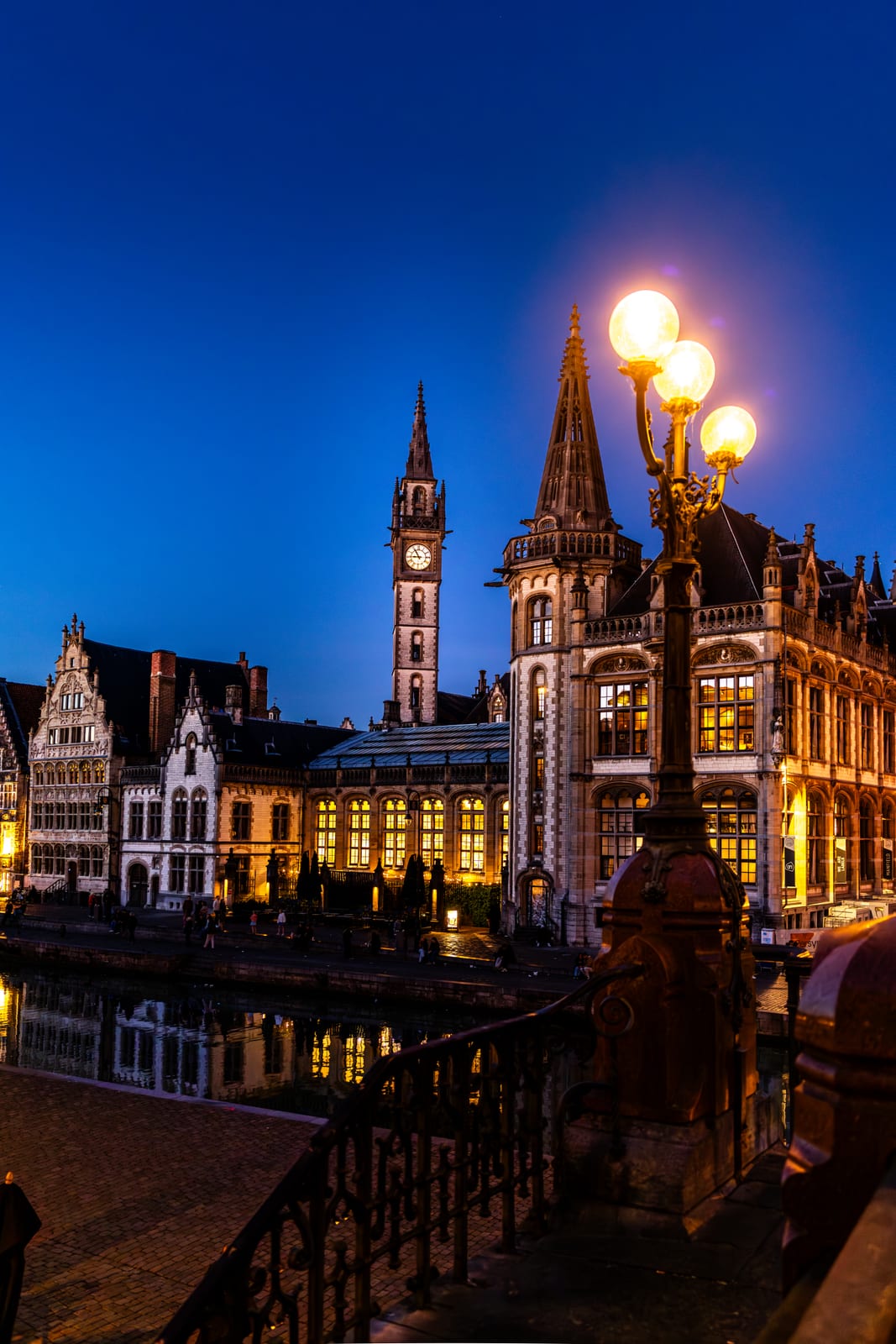 The Graslei in Ghent at blue hour, a lit street lamp in the foreground and the belfry clock tower rising behind the medieval facades.