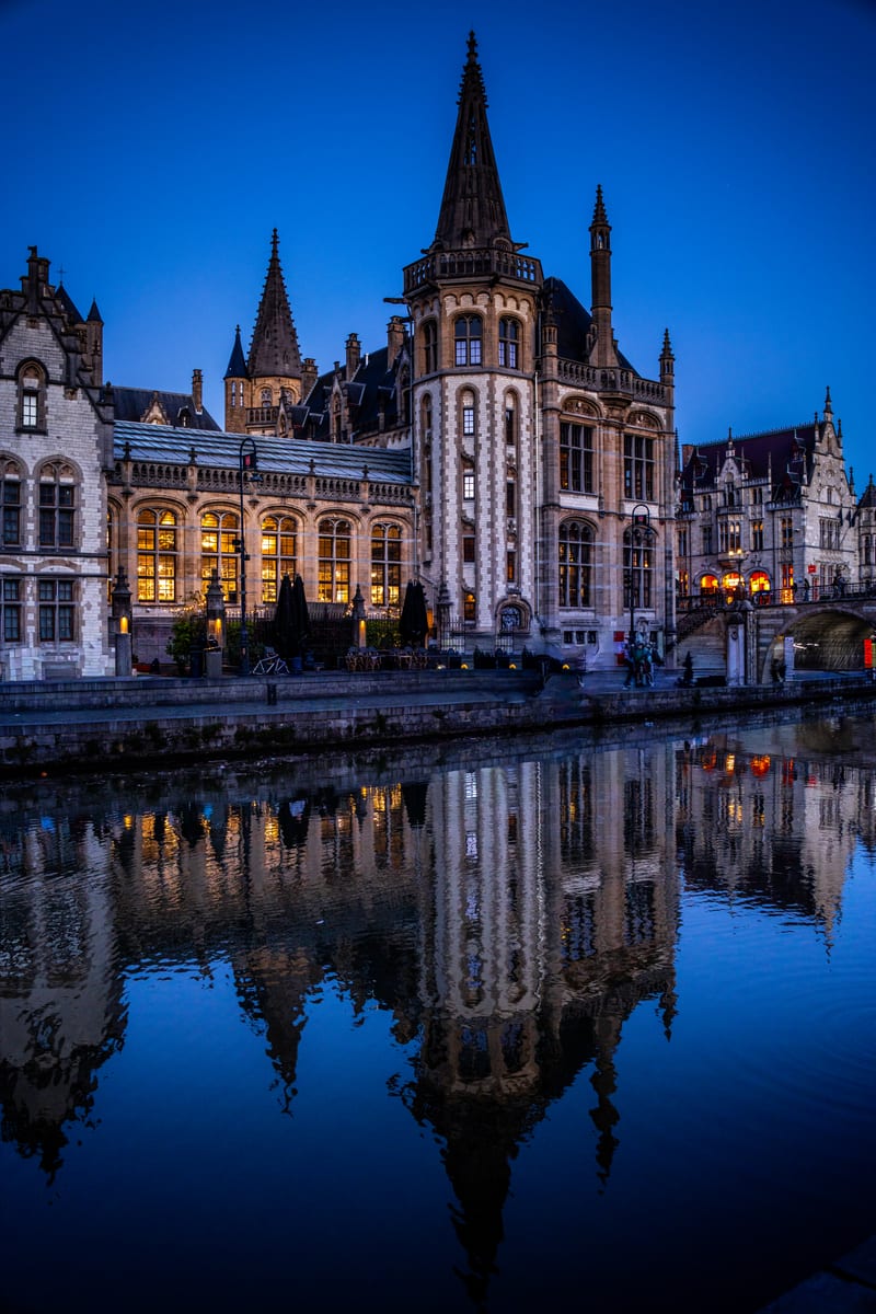 Stepped-gable guild houses along the Graslei quay in Ghent, lit from within and reflected in the still water of the Leie at blue hour.
