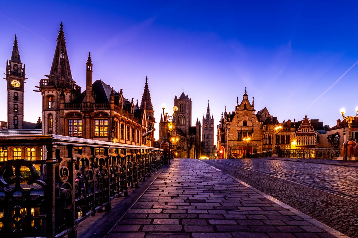 The view from Saint Michael's Bridge over Ghent before sunrise: wet cobbles in the foreground, spires and gabled houses under a deep blue sky, street lamps still lit.