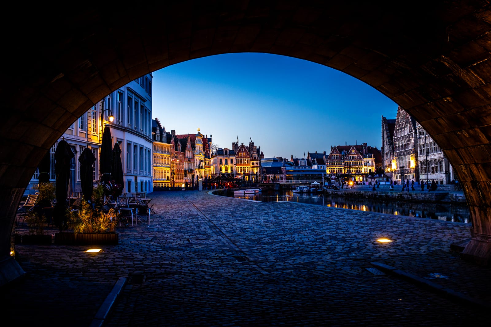 The Graslei seen through the dark arch of a stone bridge, the lit quay and its gabled houses framed inside the opening at night.