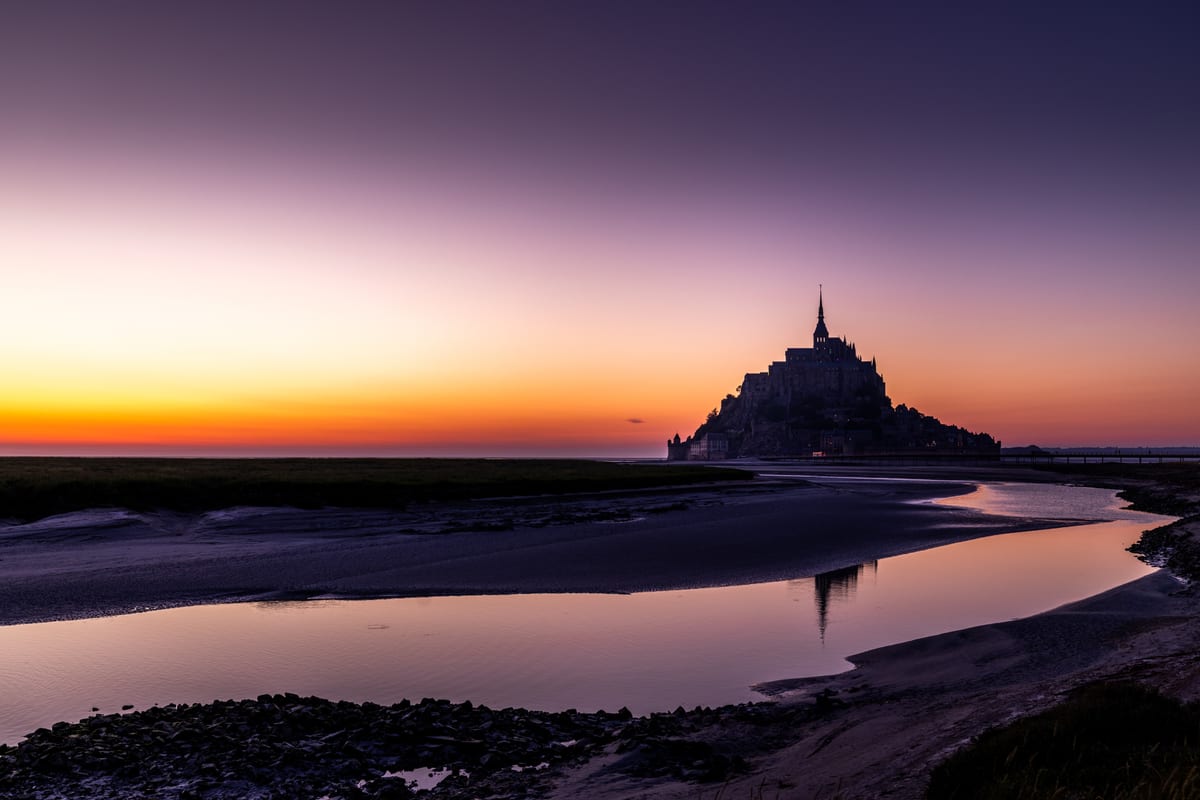 Mont Saint-Michel in silhouette after sunset, reflected in a channel of water left on the sand at low tide, the sky graded from orange to violet.