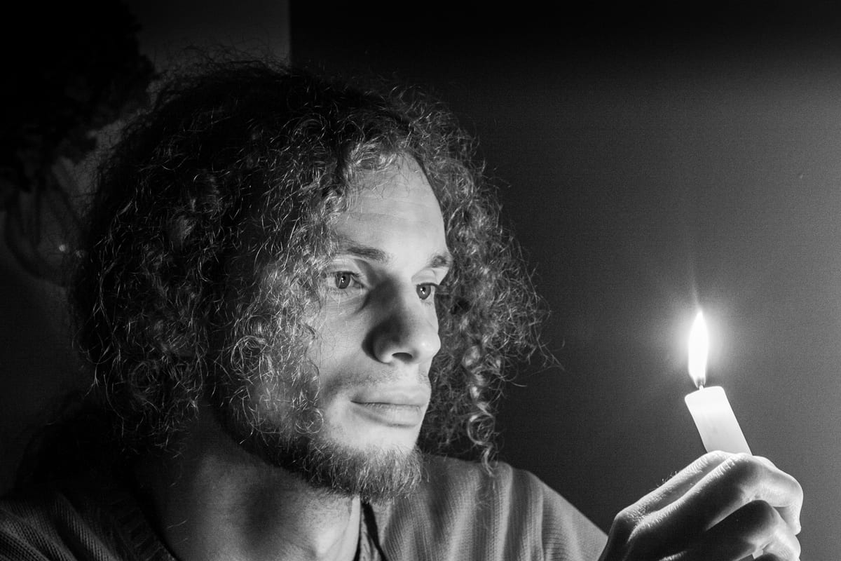 A black and white portrait of a young man with curly hair holding a lit candle, his face lit only by the flame against a dark background.