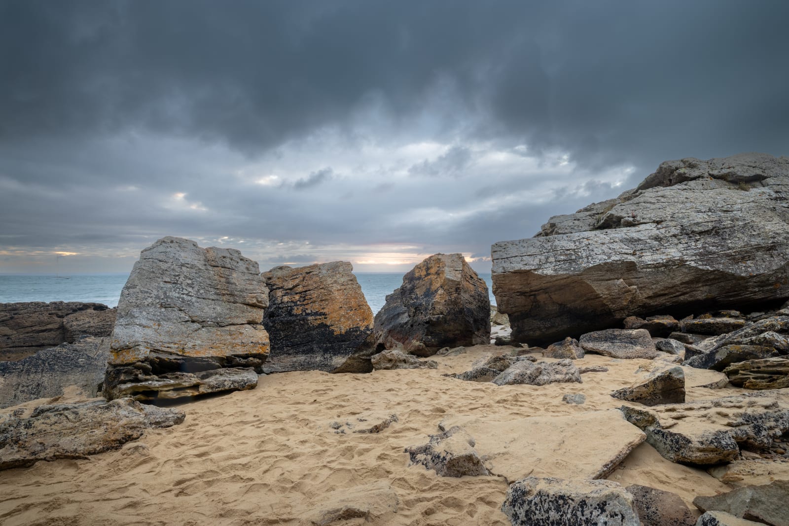 Angular pale rocks on a sand beach under a heavy grey and blue cloud bank, the sea flat behind them.