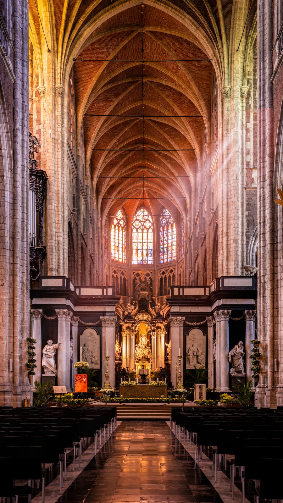 The nave of Saint Bavo's Cathedral in Ghent seen from the entrance, brick vaults rising above white stone columns towards the lit high altar.