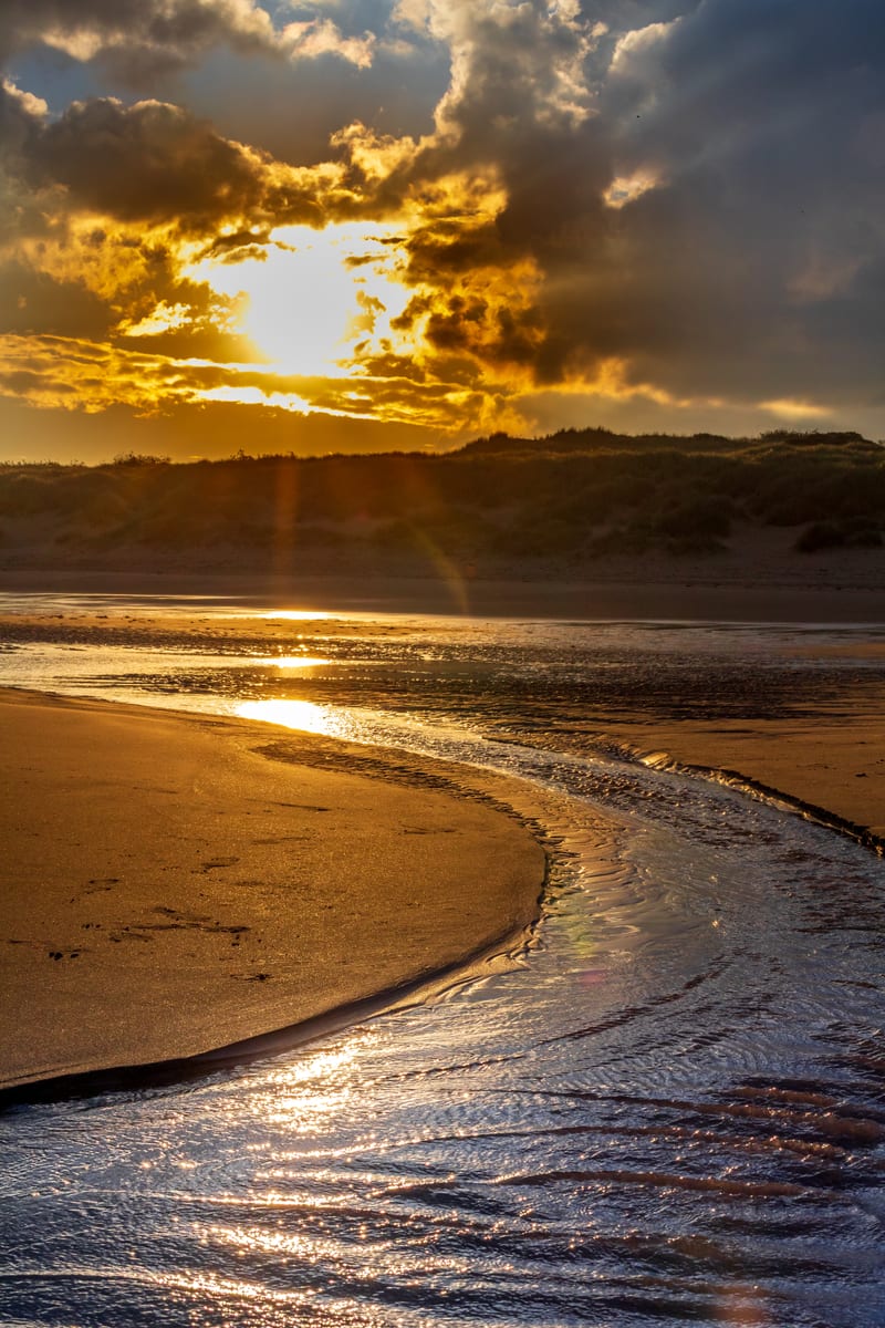 A channel of seawater winding across the beach at low tide, catching the gold of the rising sun through broken cloud.