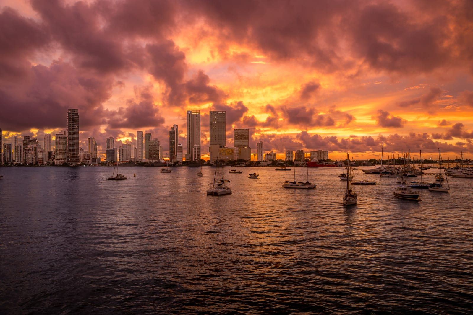 The modern skyline of Cartagena seen from the water at sunset, sailing boats moored in the bay under a sky of orange and violet cloud.