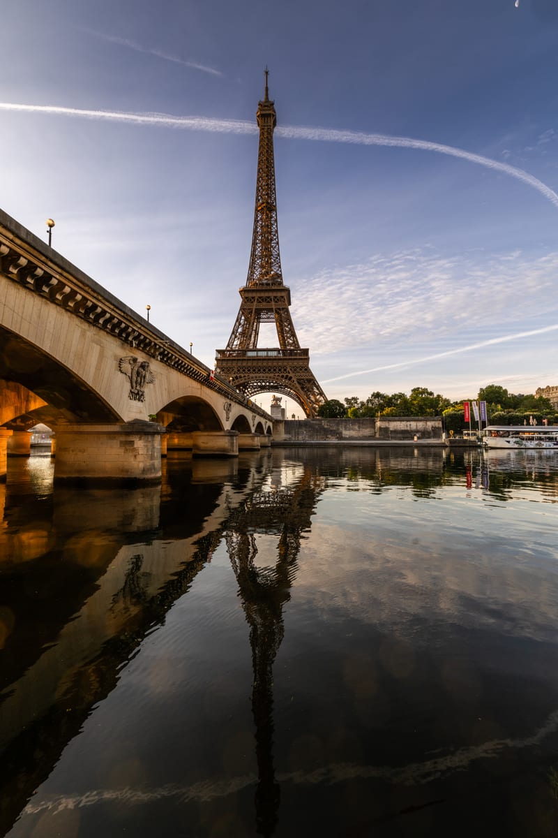 The Eiffel Tower seen from the quay beside the Pont d'Iéna in the morning, mirrored in the still water of the Seine.