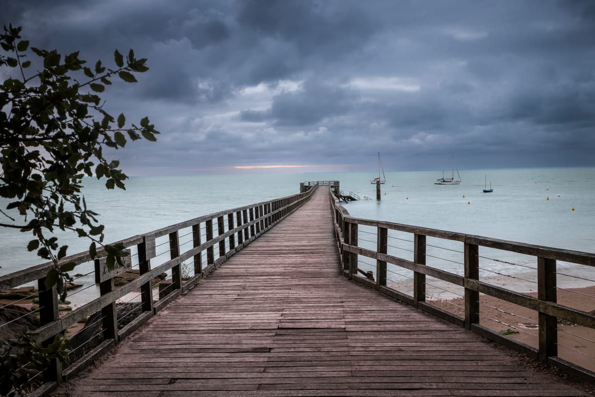 A wooden jetty running out over pale green water towards a distant boat, under a heavy grey cloud bank with a thin band of light at the horizon.